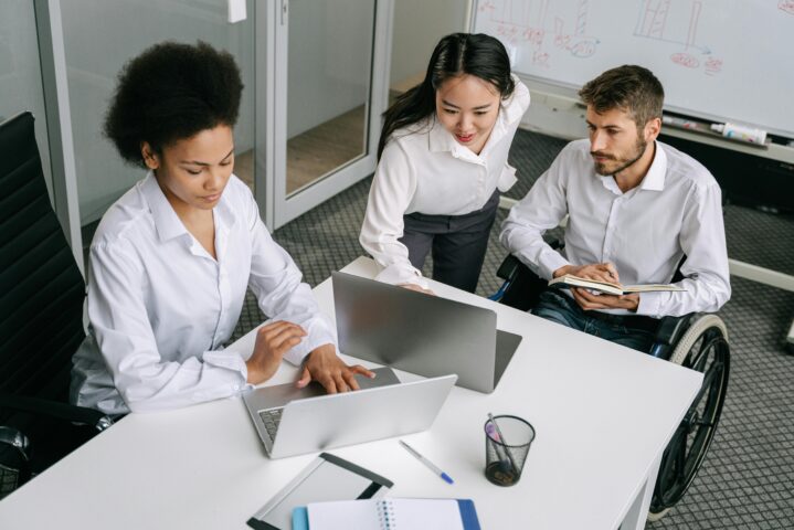 The image shows three people gathered around a desk, working together on laptops. One person, a man using a wheelchair, is taking notes in a notebook while watching the others. Another person, seated, is typing on a laptop and wearing a focused expression. The third person is standing, leaning over slightly to engage with the seated team members. Behind them, there is a whiteboard with various charts and notes, and the setting appears to be a modern office space, suggesting a collaborative and inclusive work environment.