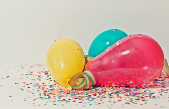 3 balloons, one pink, one yellow, and one blue, are laying on a bed of rainbow confetti.