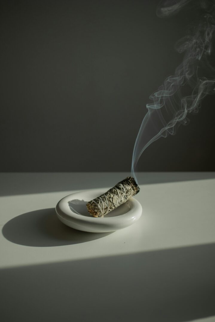 This image features a smoldering bundle of sage resting in a round, white ceramic dish. The sage bundle is tightly wrapped with twine and emits delicate, swirling trails of smoke that drift upward, creating a soft, calming atmosphere. The background is a muted, dark gray, and natural light casts soft shadows across the white surface, emphasizing the minimalistic and serene aesthetic of the scene.