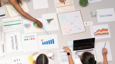 Four people sit around a large table covered with marketing and business planning materials. The table is filled with colorful charts, graphs, and documents labeled with terms like “MARKETING STRATEGY,” “TRENDS,” “ANALYSIS,” and “INSURANCE.” One person points to a Venn diagram, another writes notes in a notebook beside a laptop, a third reviews a spreadsheet, and the fourth examines a document labeled “strategy.” A potted plant, calculator, cup of coffee, and sticky notes are also visible, reflecting a collaborative and analytical work session.
