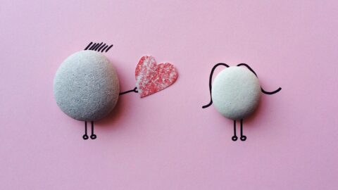On a pink background, two smooth round stones are creatively turned into simple cartoon-like characters using black line drawings for arms, legs, and hair. The stone on the left has short lines drawn on top as hair and is holding a red, heart-shaped cutout toward the stone on the right, which has curved lines for hair. The scene evokes a sweet, whimsical feeling of connection and care.