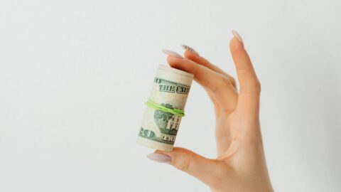 A close-up of a person’s hand holding a small roll of U.S. twenty-dollar bills secured with a green rubber band. The hand has long, manicured nails with a soft pink polish and one nail featuring black design detail. The background is a plain white wall, giving the image a clean and minimal aesthetic.