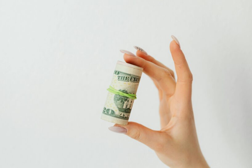 A close-up of a person’s hand holding a small roll of U.S. twenty-dollar bills secured with a green rubber band. The hand has long, manicured nails with a soft pink polish and one nail featuring black design detail. The background is a plain white wall, giving the image a clean and minimal aesthetic.