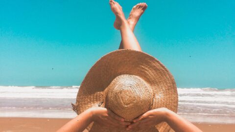 A person lies on a sandy beach with their arms stretched forward, hands resting on a large woven sunhat that covers their face. Their legs are playfully crossed in the air as they relax near the shoreline. The ocean waves roll gently in the background under a bright blue, cloudless sky.