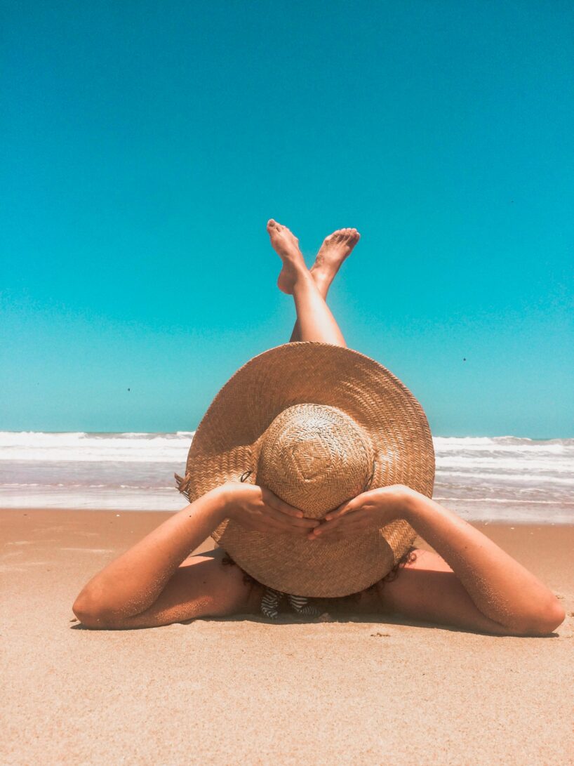 A person lies on a sandy beach with their arms stretched forward, hands resting on a large woven sunhat that covers their face. Their legs are playfully crossed in the air as they relax near the shoreline. The ocean waves roll gently in the background under a bright blue, cloudless sky.