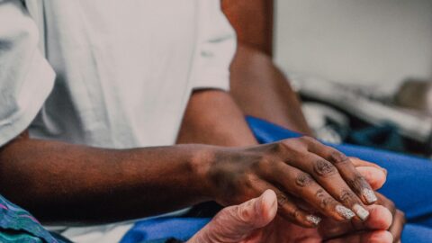 A close-up photo of a volunteer with dark skin gently holding the hands of an older adult. The volunteer is wearing a white T-shirt with the word “VOLUNTEER” printed in bold black letters. Their hand rests softly in the older adult’s open palm in a comforting, supportive gesture. The older adult is wearing a blue plaid shirt and ripped jeans. The background is softly blurred, keeping the focus on the caring interaction between the two people.