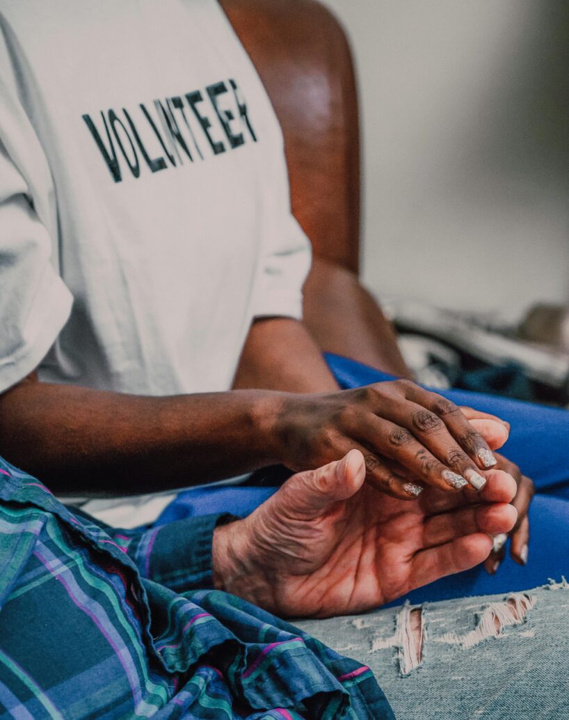 A close-up photo of a volunteer with dark skin gently holding the hands of an older adult. The volunteer is wearing a white T-shirt with the word “VOLUNTEER” printed in bold black letters. Their hand rests softly in the older adult’s open palm in a comforting, supportive gesture. The older adult is wearing a blue plaid shirt and ripped jeans. The background is softly blurred, keeping the focus on the caring interaction between the two people.