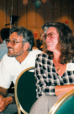 A candid photo of Networks co-founders Mike and Rosa McAllister seated side by side at an event, both smiling warmly as they look toward something off to the left. Mike has gray hair, a salt-and-pepper beard, and glasses, and wears a white button-up shirt. Rosa has long, curly brown hair and glasses, and wears a black-and-white checked shirt with a pendant necklace. Both wear name tags. Behind them, colorful balloons float near the ceiling of a warmly lit room. The photo has a soft, slightly faded look that suggests it was taken in the early years of Networks.